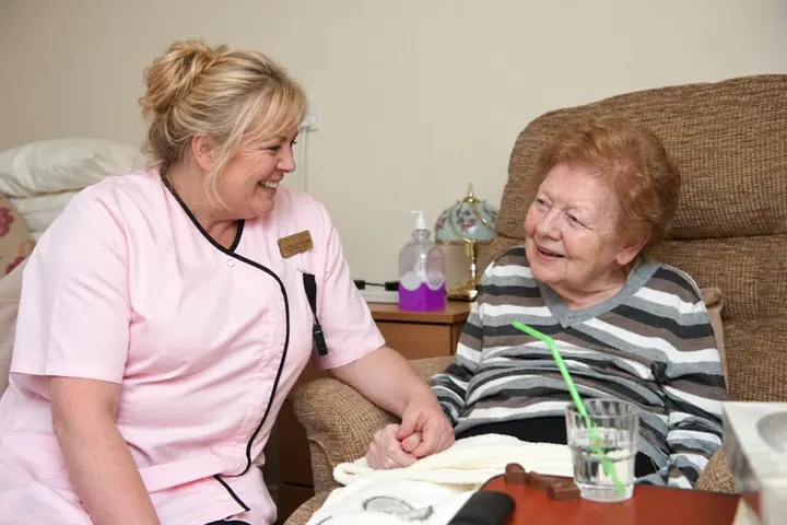 A carer in a pink tunic holding a resident&rsquo;s hand as they smile at each other beside her armchair