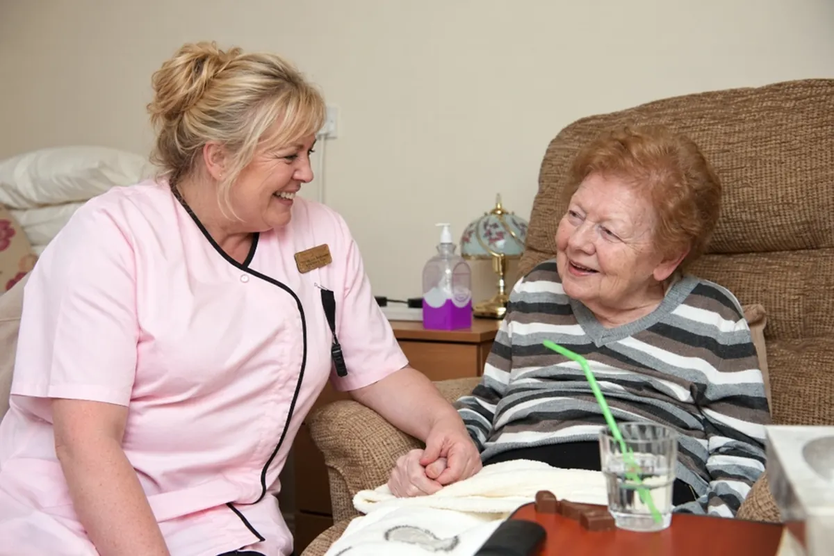 A carer in a pink tunic holding a resident&rsquo;s hand as they smile at each other beside her armchair