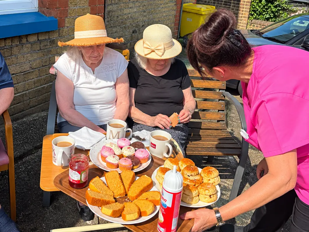 Two residents in straw sun hats on a bench beside the house as a carer brings a tray of tea, cakes and scones